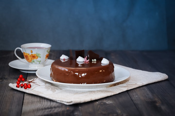 On a dark tabletop, on a linen napkin on a white dish, there is a round chocolate cake, next to a white cup is dark tea and next to the round berries