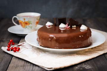 On a dark tabletop, on a linen napkin on a white dish, there is a round chocolate cake, next to a white cup is dark tea and next to the round berries