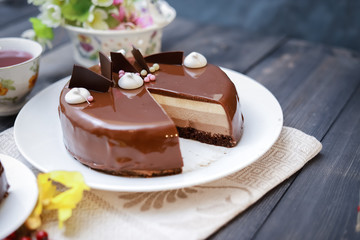 On a dark tabletop on a linen napkin, a tea composition: on a saucer a piece of cake made of three chocolate layers of different colors souffle, covered with glossy chocolate