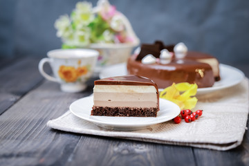 On a dark tabletop on a linen napkin, a tea composition: on a saucer a piece of cake made of three chocolate layers of different colors souffle, covered with glossy chocolate