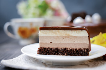 On a dark tabletop on a linen napkin, a tea composition: on a saucer a piece of cake made of three chocolate layers of different colors souffle, covered with glossy chocolate