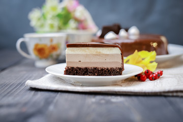 On a dark tabletop on a linen napkin, a tea composition: on a saucer a piece of cake made of three chocolate layers of different colors souffle, covered with glossy chocolate