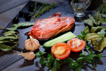 A piece of raw beef is laid out on a dark wooden tabletop on a graphite stone tile, next to a sauceboat with butter, peas, peppermint, laurel leaves, thyme, olives, garlic
