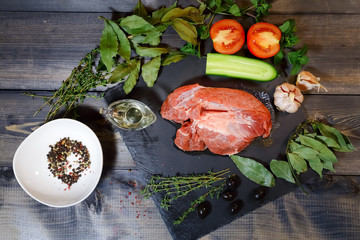 A piece of raw beef is laid out on a dark wooden tabletop on a graphite stone tile, next to a sauceboat with butter, peas, peppermint, laurel leaves, thyme, olives, garlic