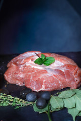 On a dark countertop, a piece of raw beef meat, decorated with thyme and chestnut leaf, black large olives and a head of garlic