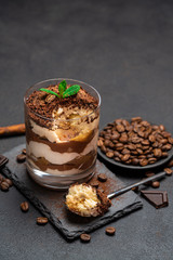 Classic tiramisu dessert in a glass cup and pieces of chocolate on stone cutting board on dark concrete background