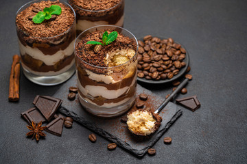 Classic tiramisu dessert in a glass cup and pieces of chocolate on stone cutting board on dark concrete background