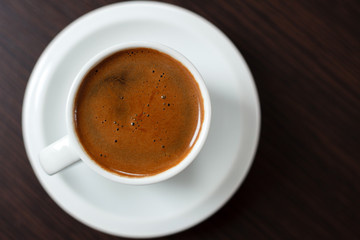 Close-up of single white cup of coffee on dark wooden table. High angle view, flat-lay. Selective focus on coffee surface