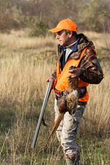 A man with a gun in his hands and an orange vest on a pheasant hunt in a wooded area in cloudy weather. A hunter with a pheasant in his hands.