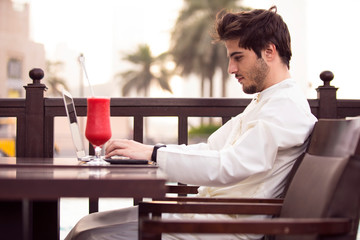 Young bearded businessman wearing Kandoura, sitting at a table in cafe and uses a laptop. Nearby smartphone and cup of juice. Man checks email on computer. Freelancer working outside office