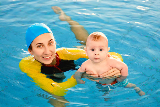 Young mother aged 30 years and baby son of 5 months are in pool. Smiling woman with beautiful snow-white smile is dressed in fashionable swimming suit swimsuit and cap. Girl teaches boy to swim.