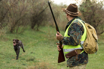 Fototapeta premium Hunting period, autumn season open. A hunter with a gun in his hands in hunting clothes in the autumn forest in search of a trophy. A man stands with weapons and hunting dogs tracking down the game.