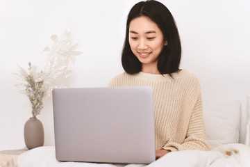 Beautiful woman freelancer Asian appearance with dark hair hairstyle Bob wears sweater has an attractive white smile works at laptop, writes post in blog about personal life career dreams of traveling