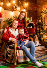 Happy family by the Christmas tree. Family goals. Baby is opening a christmas gift. Lovely kissing parents on the background. Father mother with his child on hands.