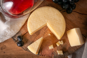 Caciotta cheese on a round wooden board. View from above. Nearby are several sliced ​​pieces of cheese and black grapes. In the background is a glass of red wine. Background gray linen fabric