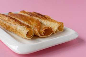 Fried pancakes roll into a tube on a white plate with a pink background.