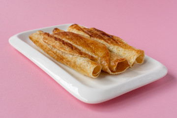 Fried pancakes roll into a tube on a white plate with a pink background.