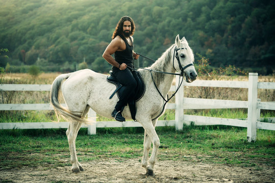 Full Length Of Young Handsome Man Sitting On His White Stallion At The Country Side. Man Equestrian On His Horse Riding At Nature. Attractive Man Sitting On White Horse On The Ranch In Autumn.