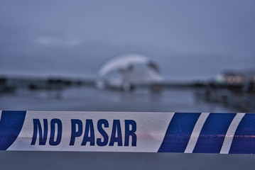 Outdoor foreground warning of not passing an object out of focus in the background on a day of strong, gray and sad winds. White and blue plastic tape. Text in Spanish. NO PASAR "No passing zone"