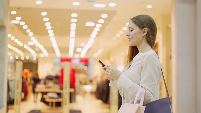 Tracking slow motion waist up shot of smiling young woman walking with shopping bags in mall and text messaging on cell phone after successful shopping