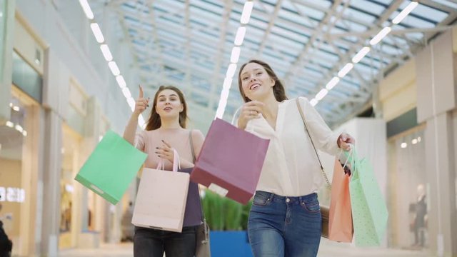 Two excited young women with shopping bags running in slow motion along mall on Black Friday hurrying to make purchase and pointing on stores