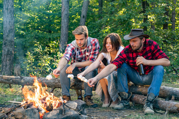 Friends enjoy weekend barbecue in forest. Company friends picnic or barbecue roasting marshmallows near bonfire. Group of backpackers relaxing near campfire. Roasting marshmallows barbecue.