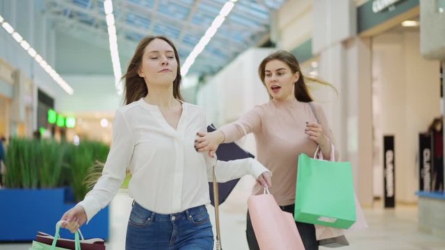 Medium slow motion shot of two excited young women with shopping bags running along mall hurrying to make purchases on Black Friday. Woman pushing off her rival to be first in store