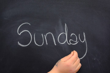 Obraz premium Woman's hand writing the day of the week on a blackboard with white chalk, sunday