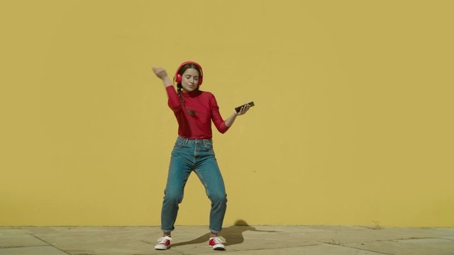 Young Happy Latin Woman Makes Dancing Cool Moves Wearing A Red Shirt And Jeans And Red Converse Shoes With A Yellow Background Wall On A Sunny Day Wide-angle Shot In Slow Motion