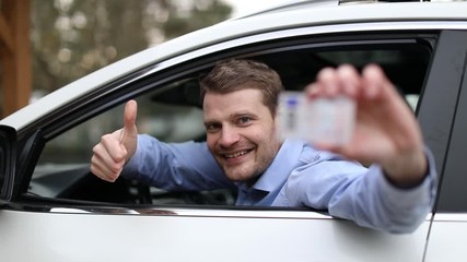 young happy man sitting in the car and showing his new driver license with thumb up