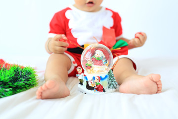 Close-up little baby boy in Santa costume playing snow globe sitting on bed over white background. Merry Christmas and Happy New Year Concept.