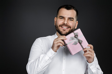 portrait of smiling handsome bearded man in white shirt holding gift box and looking at camera isolated on black background