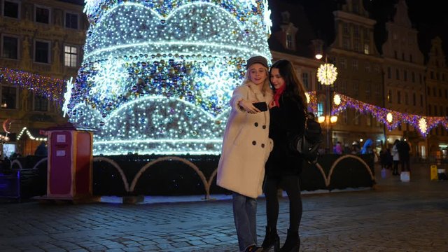 Two pretty young girlfriends women take selfie picture near christmas tree in night city center in Poland