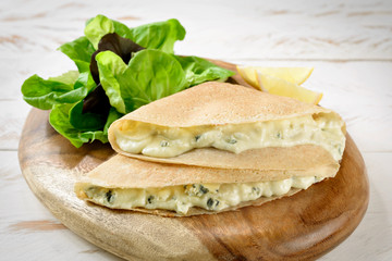 Crepe with gorgonzola cheese, with salad aside. On a cutting board on a wooden table.
