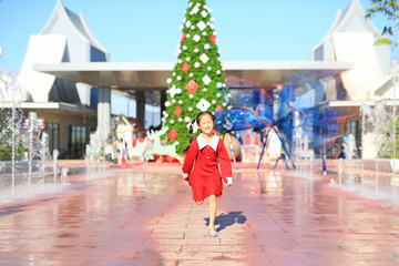 Smiling little Asian girl in red dress running around the big christmas tree decorative for the happy new year and merry christmas festival.