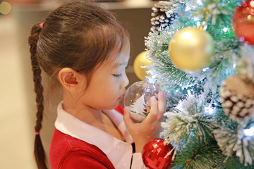 Cute Asian little girl in red dress holding and looking decorative ball of christmas tree in the winter season and happy new year.