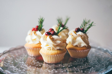 Beautiful christmas cupcakes with curd cream, cranberries and spruce branches on top on the glass table against a white wall.