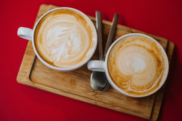 Top view image cup of hot cappuccino with latte art at cafe on red table and rustic wooden tray. Close up cups strong coffee with beautiful foam stand on red background