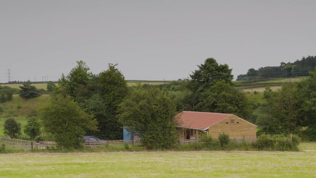 A still medium shot of a little house with a car parked outside with trees surrounding and a car driving off in the distance
