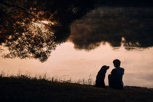 woman and dog silhouettes view, sitting by the river together in summer