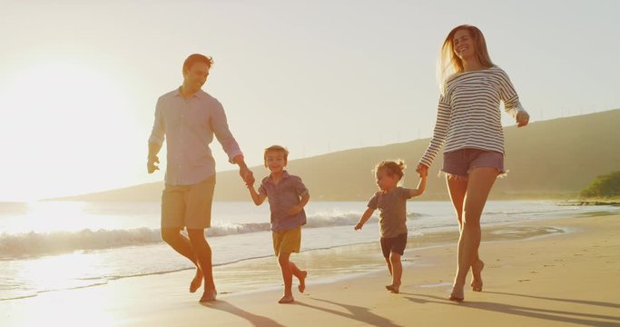Happy smiling toddler boys holding hands with their parents running together on the beach, young joyful family having fun on the beach at sunset