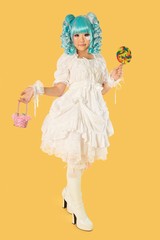Full length portrait of young woman dressed as a doll holding lollipop over yellow background