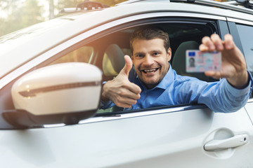 young smiling man sitting in the car and showing his new driver license with thumb up sign out of...