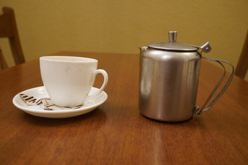 Old dirty used aluminum tea pot kettle with dirty used white tea mug. Antique Aluminum Kettle with tea cup and empty used sugar sachets. Dirty coffee and tea cup half empty on saucer on table.