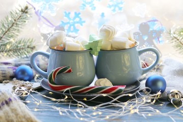 Two cups with a drink, marshmallows, caramel sticks, Christmas decor, illuminations on a wooden surface on a window background,  winter holidays, blue color