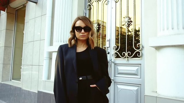 Fashionable, beautiful brunette girl with wavy hair in a black men's suit and glasses walks around the city