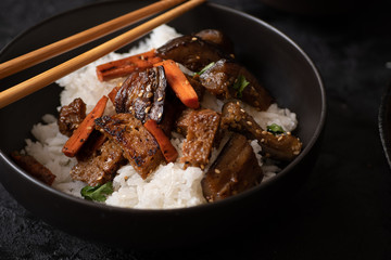 Vegan Korean seitan, carrot and eggplant fry over rice with kimchi in a black bowl