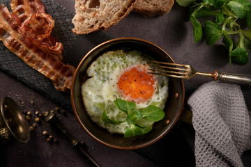 Fried egg in a frying pan with bacon and bread for breakfast on dark background, decorated valerian grass, flat lay, vintage style