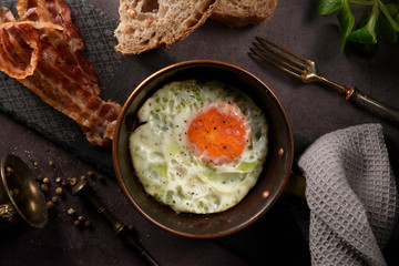 Fried egg in a frying pan with bacon and bread for breakfast on dark background, decorated valerian...