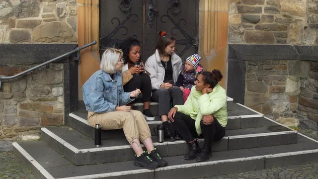 Multiracial Group of Young Girlfriends and Cute Little Girl Having Good Time while Sitting on the Steps of Ancient European Building. They Talking and Smiling. Friendship, Lifestyle and People Concept